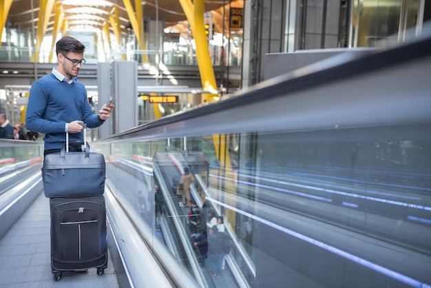Молодой человек на эскалаторе в аэропорту, используя свой мобильный телефон с багажом улыбается