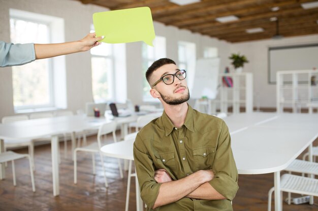 Young man in eyeglasses and shirt dreamily looking aside while woman hand holding paper shape of message under his head at work in modern cozy office