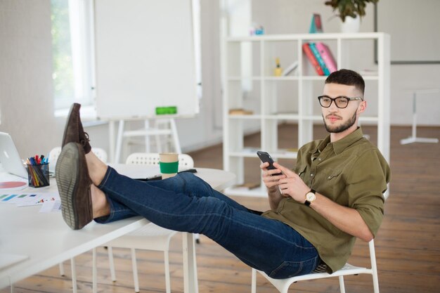 仕事でテーブルの上に足で椅子に座っている間携帯電話を保持している眼鏡とシャツの若い男現代のオフィスで夢のように脇を探している創造的なビジネスマン