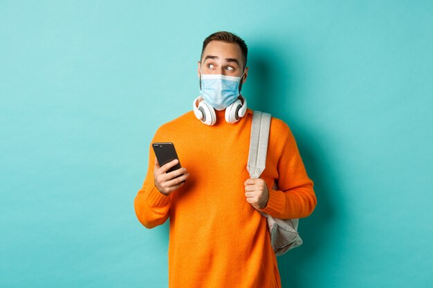 Young man in face mask using mobile phone, holding backpack, staring left amazed