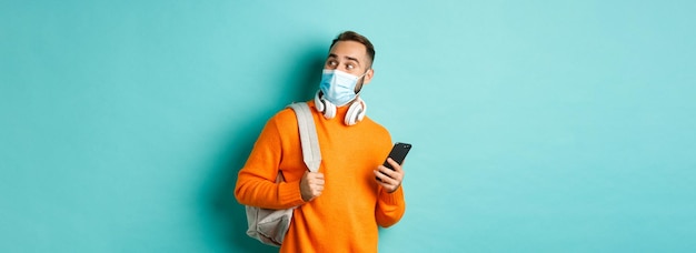 Free photo young man in face mask using mobile phone holding backpack staring right amazed standing against lig