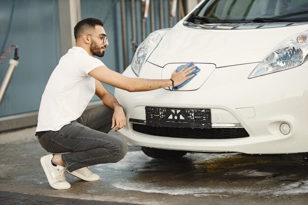 洗車場で洗車した後、ラグで車を拭くファッション服を着た青年。白いTシャツとジーンズを着ている男