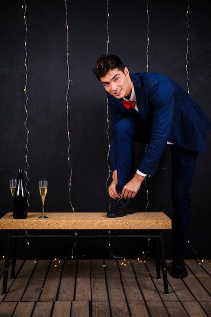 Young man fastening shoe laces near glass and bottle of champagne 