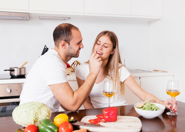 Foto gratuita giovane che alimenta il bellpepper a sua moglie con i bicchieri di vino sulla tavola di legno