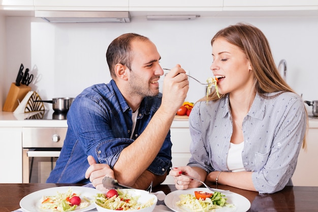 Молодой человек кормит салат своей жене, сидящей на кухне