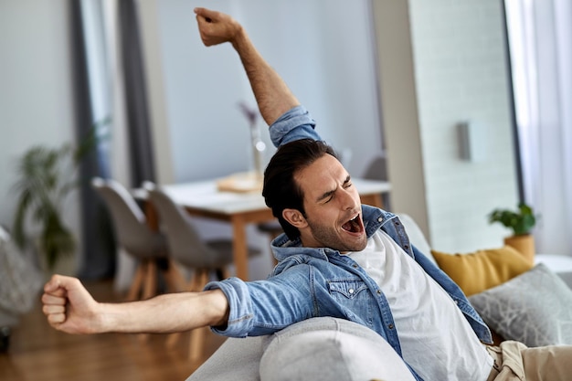 Young man feeling tired and yawning while sitting on the sofa at home