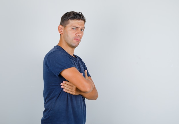 Young man folding hands on chest with frowning eyebrows in dark blue t-shirt and looking confident.