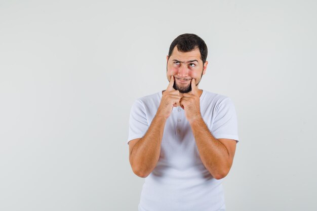 Young man forcing a smile on face in white t-shirt and looking gloomy , front view.