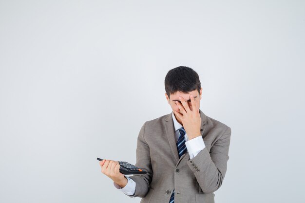 Young man in formal suit holding calculator, covering face with hand and looking annoyed