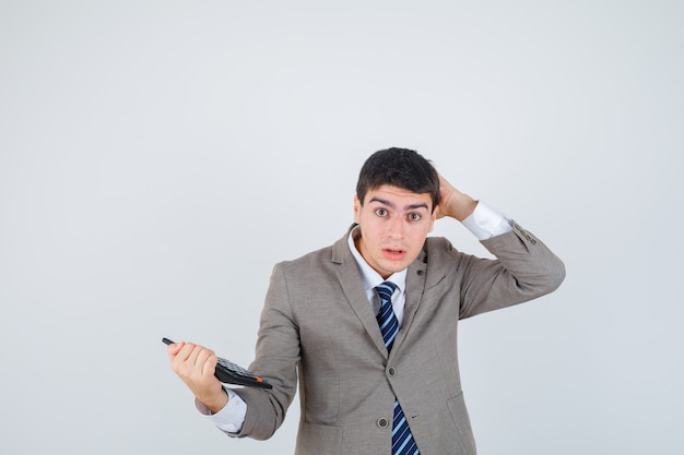 Young man in formal suit holding calculator, scratching head and looking pensive