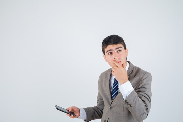 Young man in formal suit holding phone, putting hand on chin, thinking about something