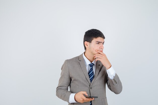 Young man in formal suit holding phone, putting hand on chin, thinking about something