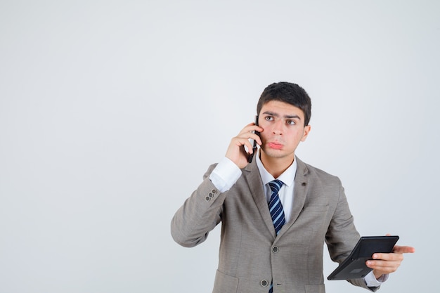 Young man in formal suit talking to phone, holding calculator, thinking about something