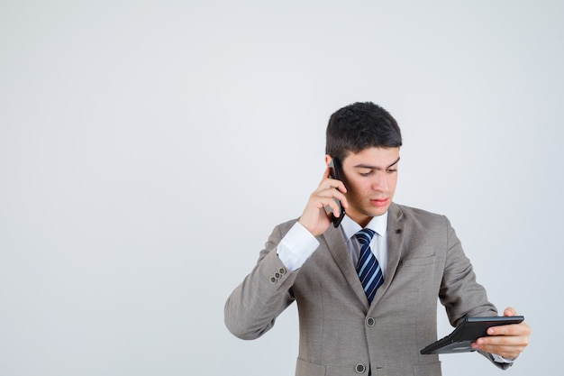 Young man in formal suit talking to phone, looking at calculator