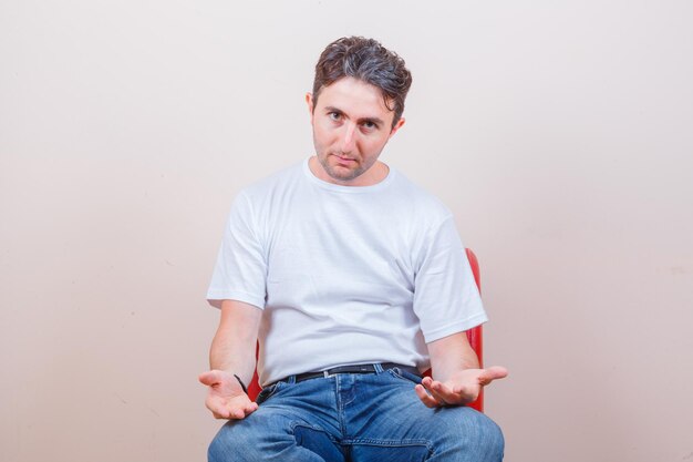 Young man gesturing while sitting on chair in t-shirt, jeans and looking confused