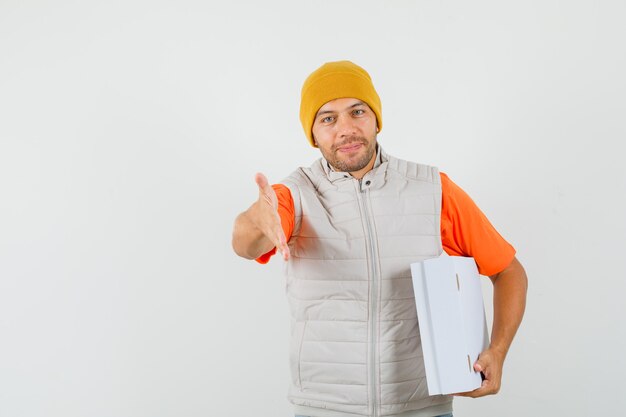 Young man giving hand for handshake, holding cardboard box in t-shirt, jacket, hat and looking gentle. front view.