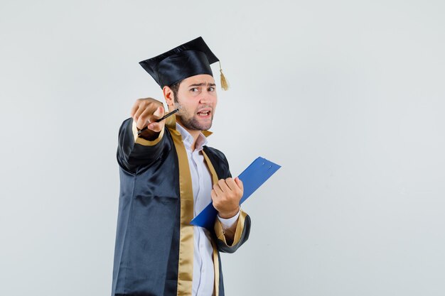 Young man in graduate uniform holding clipboard, pointing at camera and looking angry , front view.