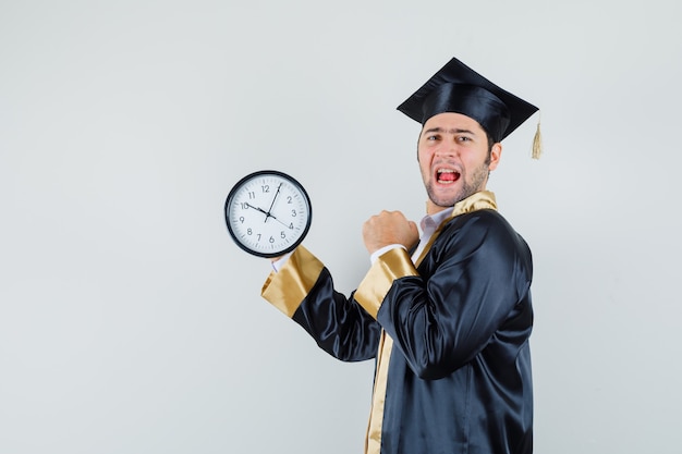 Young man in graduate uniform holding wall clock and looking happy .