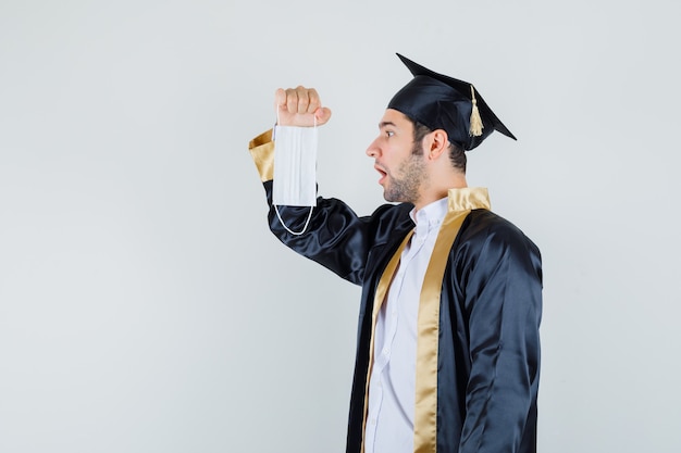 Young man in graduate uniform looking at medical mask and looking focused , front view.