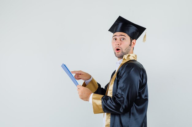 Young man in graduate uniform looking over notes on clipboard and looking wondered .