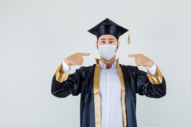 Young man in graduate uniform pointing at his medical mask , front view.