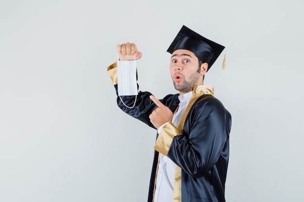 Free photo young man in graduate uniform pointing at medical mask and looking surprised.