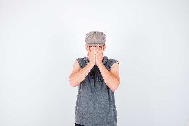 Young man in gray t-shirt and cap covering face with hands and looking annoyed