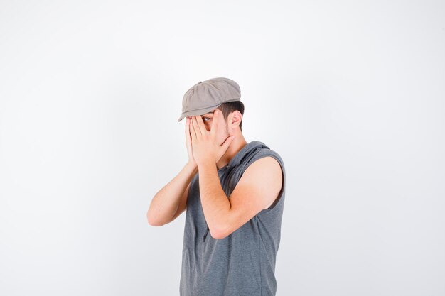 Молодой человек в серой футболке и кепке закрывает лицо руками, смотрит сквозь пальцы и выглядит раздраженным