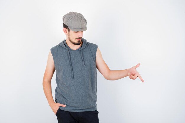 Молодой человек в серой футболке и кепке указывает вниз указательным пальцем, держит руку в кармане и выглядит серьезным