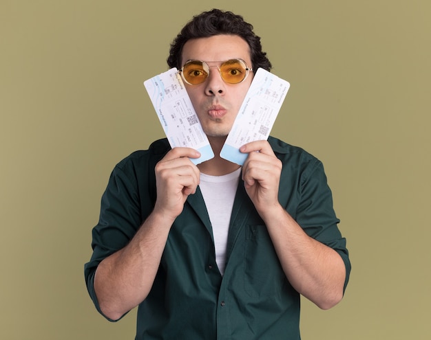 Молодой человек в зеленой рубашке в очках держит билеты на самолет и смотрит в замешательство, стоя над зеленой стеной