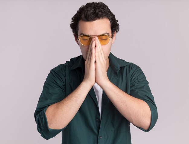 Young man in green shirt wearing glasses holding palms together tired and bored standing over white wall