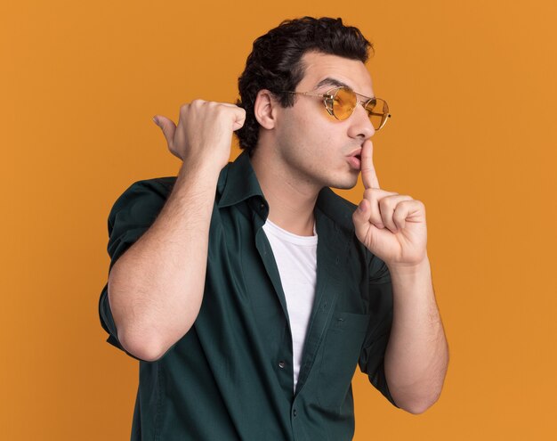 Young man in green shirt wearing glasses looking aside making silence gesture with finger on lips standing over orange wall