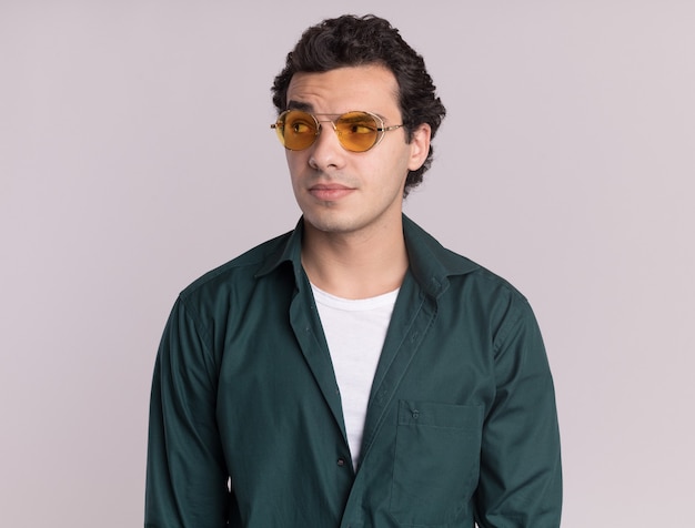 Young man in green shirt wearing glasses looking aside with skeptic expression standing over white wall