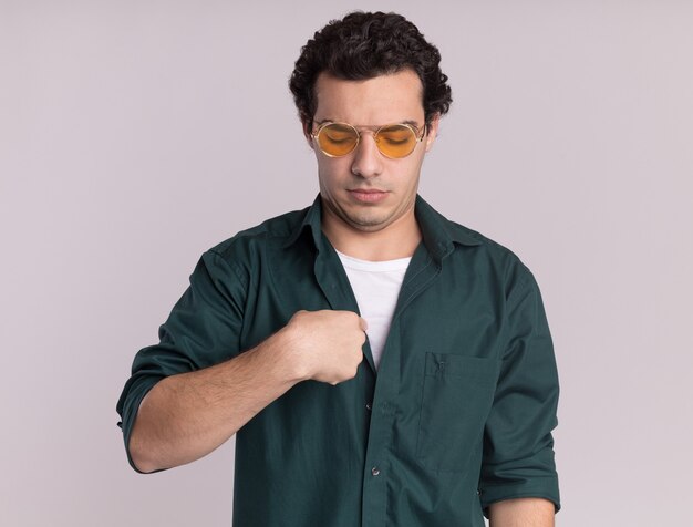 Young man in green shirt wearing glasses looking confused pointing at himself standing over white wall