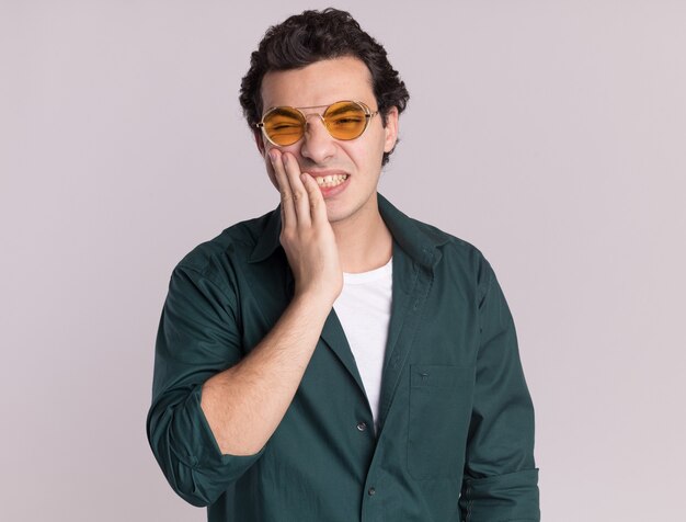 Young man in green shirt wearing glasses looking confused with hand on his mouth standing over white wall