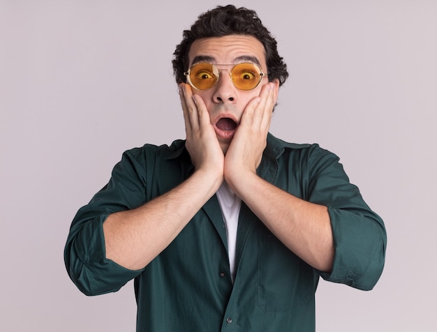 Giovane uomo in camicia verde con gli occhiali guardando davanti stupito e sorpreso in piedi sopra il muro bianco