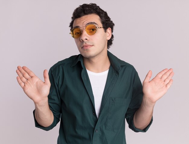 Young man in green shirt wearing glasses looking at front confused with arms raised having no answer standing over white wall