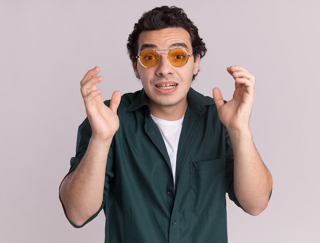 Young man in green shirt wearing glasses looking at front confused with raised arms standing over white wall