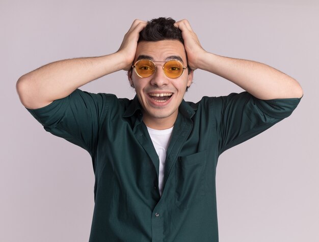 Young man in green shirt wearing glasses looking at front happy and excited with hands on his head standing over white wall