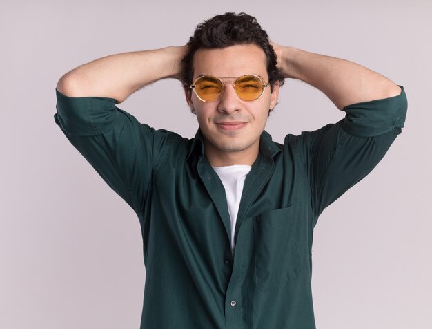 Giovane uomo in camicia verde con gli occhiali guardando davanti felice e positivo con le mani dietro la testa in piedi sul muro bianco