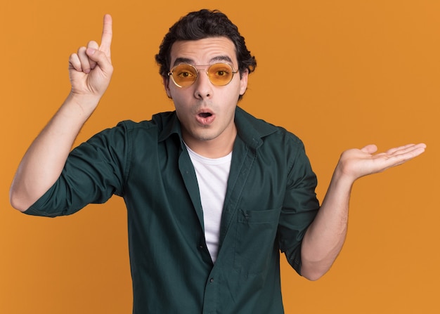 Giovane uomo in camicia verde con gli occhiali guardando la parte anteriore sorpreso indicando con il dito indice in su che presenta con lo spazio della copia del braccio che sta sopra la parete arancione