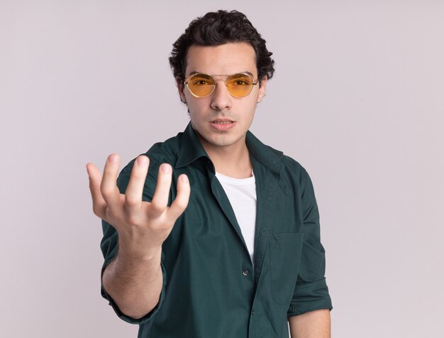 Young man in green shirt wearing glasses looking at front with angry face with arm raised standing over white wall