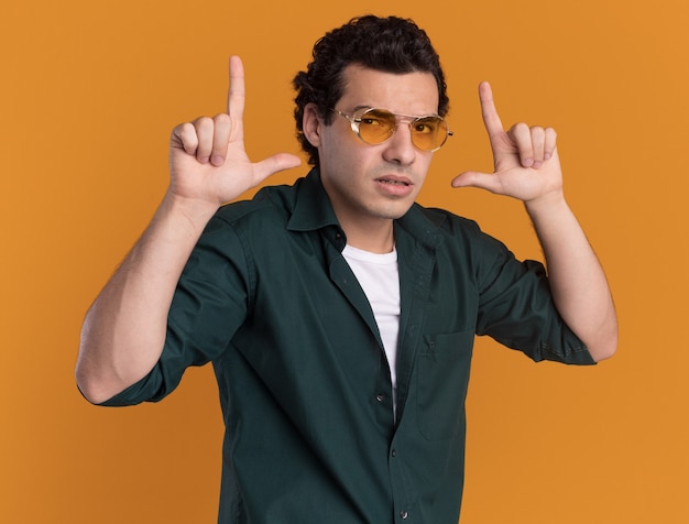 Молодой человек в зеленой рубашке в очках смотрит вперед с уверенным выражением лица, делая жест пистолета с пальцами, стоящими над оранжевой стеной