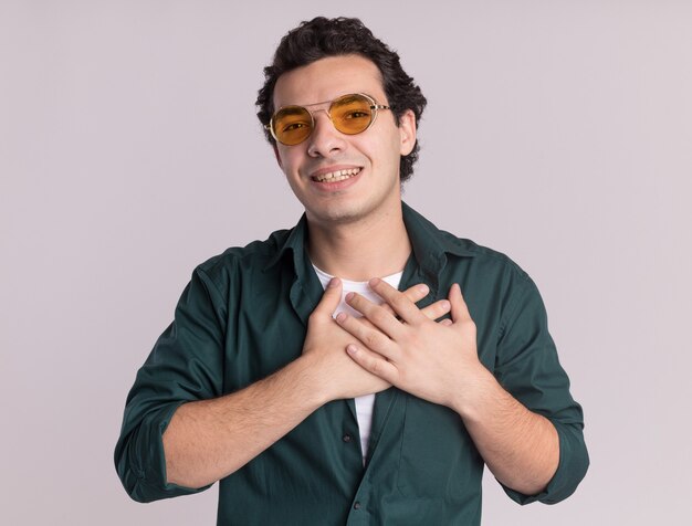 Young man in green shirt wearing glasses looking at front with hands on his chest feeling thankful standing over white wall