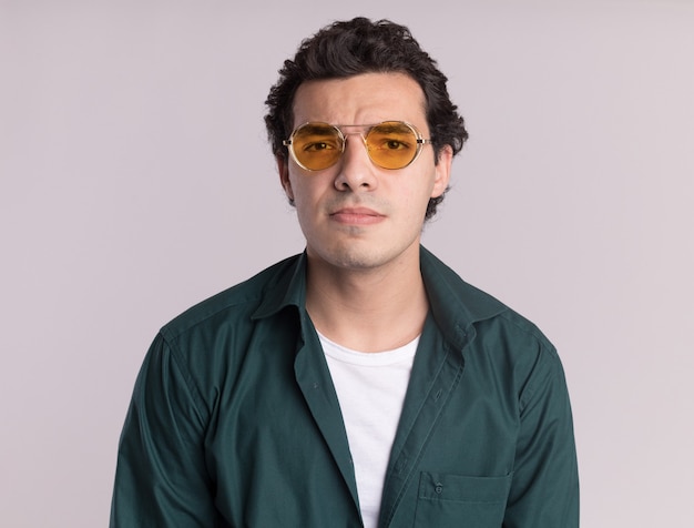 Young man in green shirt wearing glasses looking at front with sad expression standing over white wall