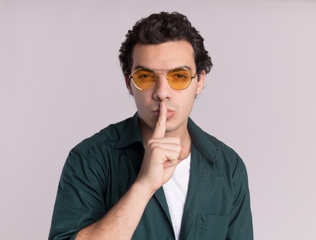 Young man in green shirt wearing glasses looking at front with serious face with finger on lips silence gesture standing over white wall