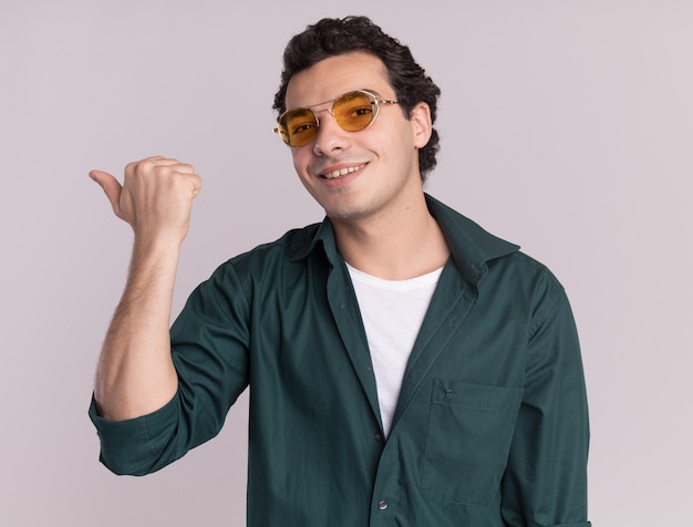 Young man in green shirt wearing glasses looking at front with smile on face pointing with thumb back standing over white wall