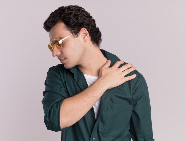 Young man in green shirt wearing glasses looking unwell touching shoulder feeling pain standing over white wall