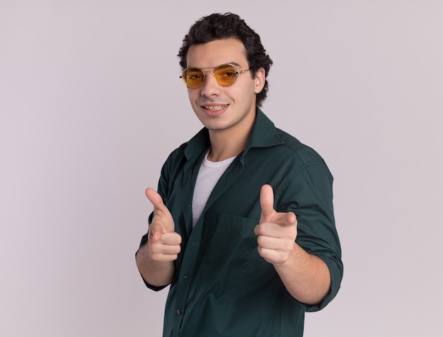 Молодой человек в зеленой рубашке в очках, указывая указательными пальцами вперед, счастливым и позитивным положением над белой стеной