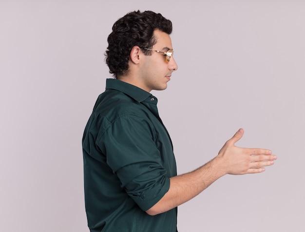 Young man in green shirt wearing glasses standing sideways offering hand greeting over white wall